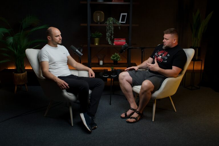 Two men sit in beige armchairs facing each other, speaking into microphones in a podcast studio setting.